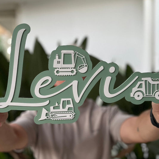 Person holding a decorative nameplate with 'Levi' and construction vehicle illustrations.