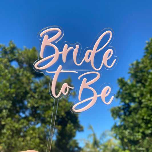 Acrylic 'Bride to Be' cake topper held against a blue sky with trees.