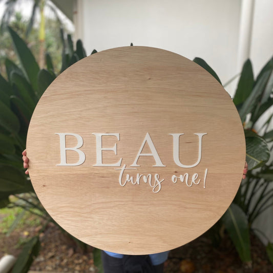 Round wooden sign with 'BEAU turns one!' text held by a person in front of plants.
